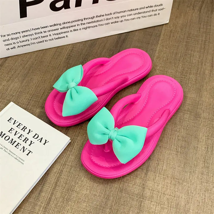 Bright pink flip-flops with large, mint green bow embellishments featuring a gold teddy bear charm, shown from a front view on a wooden surface next to a book.