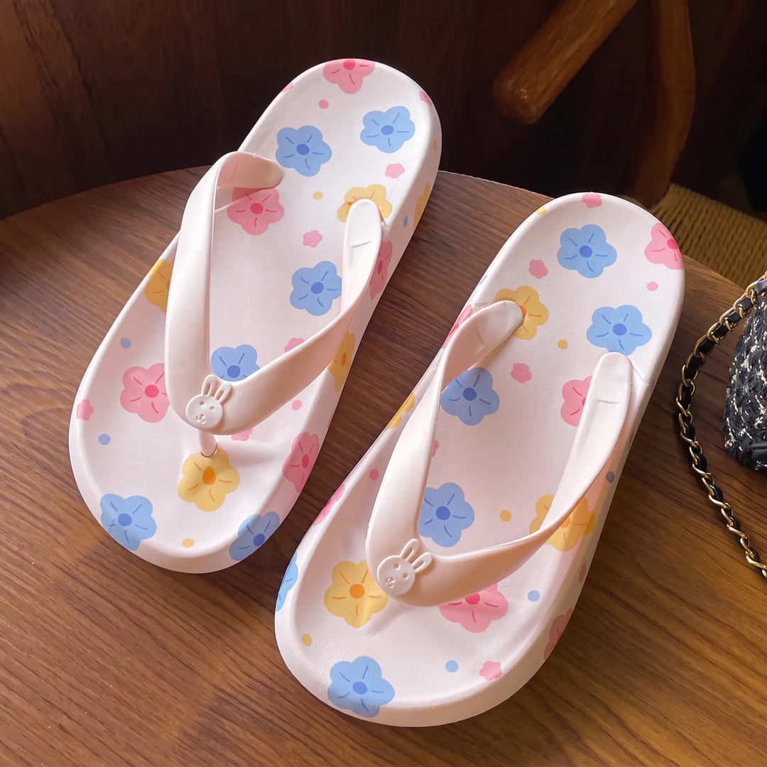 Close-up of two pink platform flip-flop sandals featuring a playful floral print and a cute bunny charm on the toe post, placed on a wooden surface.