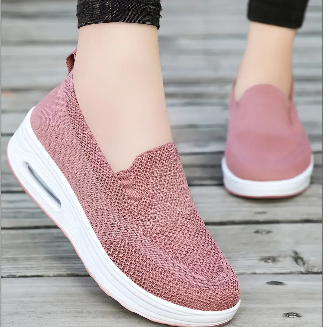 Pair of pink breathable knit slip-on sneakers with a white platform sole and visible air cushion in the heel, worn on feet against a wooden background.
