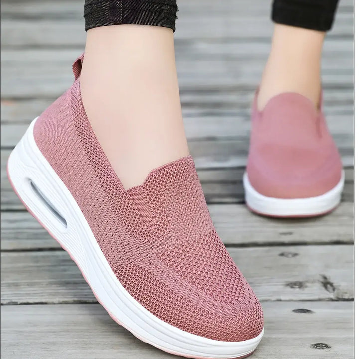 Pair of pink breathable knit slip-on sneakers with a white platform sole and visible air cushion in the heel, worn on feet against a wooden background.