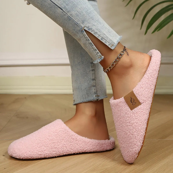 A person wearing soft pink plush clog slippers with white interior lining and a small faux leather accent tag on the outer side, set against a wooden floor.