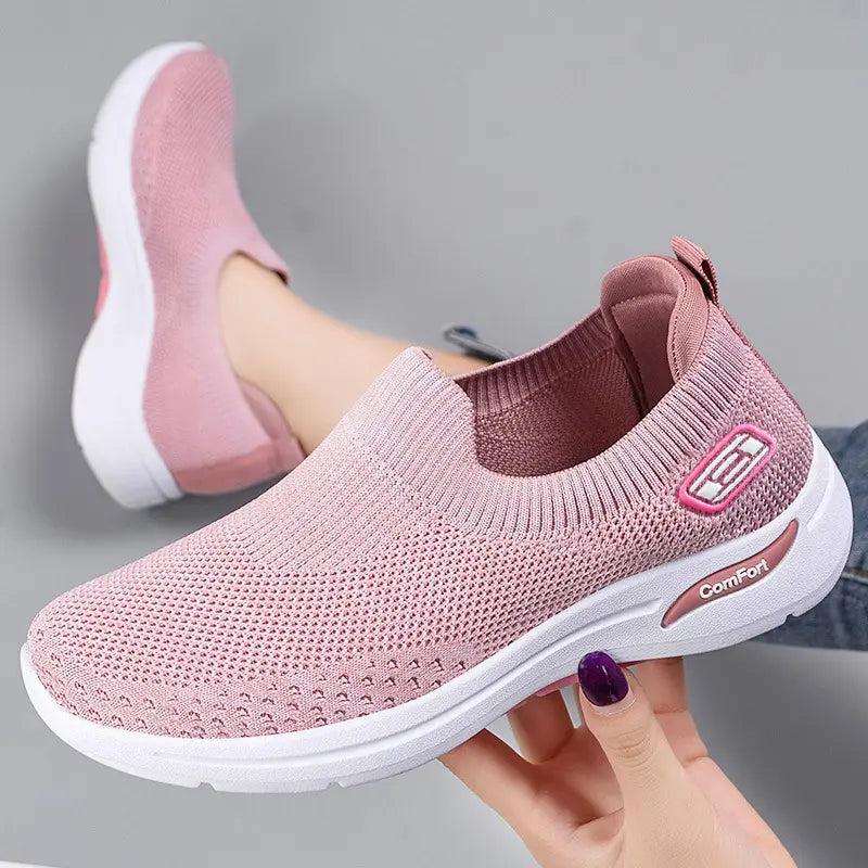 Three-quarter view of a pink breathable knit slip-on sneaker for women, featuring a white sole with 'Comfort' text and a subtle side logo. A foot wearing a matching shoe is visible in the background.