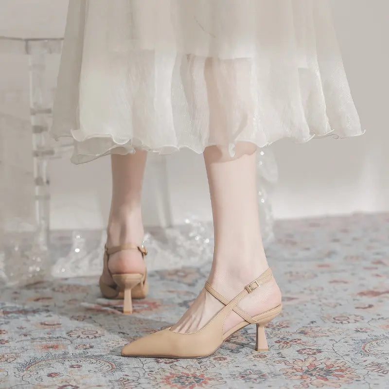 A pair of warm apricot pointed-toe slingback heels with thin adjustable ankle straps, worn by a person standing on a decorative rug, with a sheer white dress hem visible.