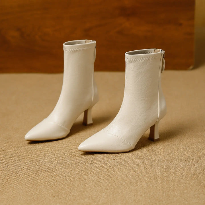 A pair of creamy ivory pointed toe ankle boots with stiletto heels and back zipper closure, shown from a three-quarter angle on a textured studio floor.