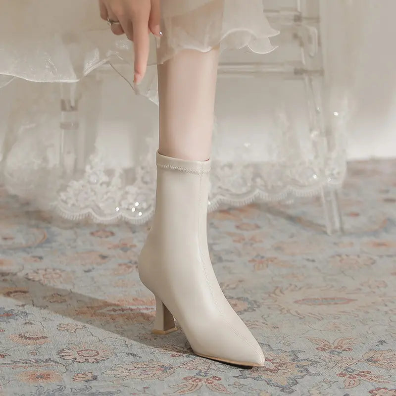 A leg wearing a chic creamy off-white pointed-toe stiletto boot, styled with an elegant white dress. Features a mid-calf shaft and sculptural flared heel.