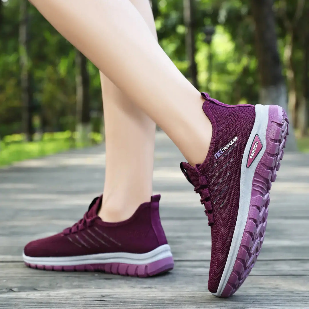 Lifestyle image of purple lightweight breathable knit sneakers worn on feet walking on a wooden path, showcasing side detail with &