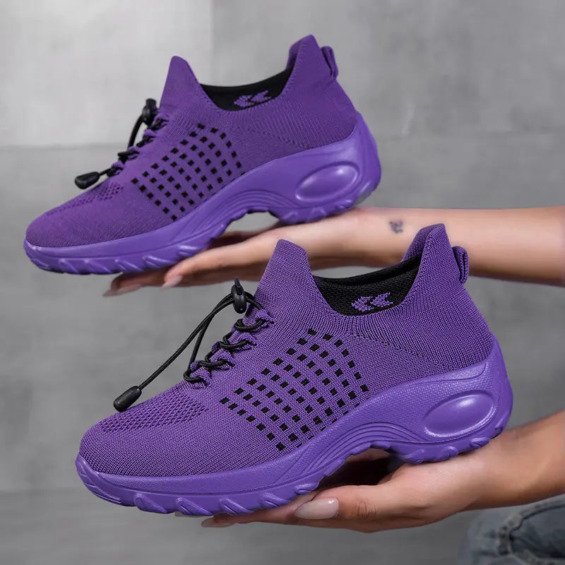 Vibrant purple women's knit sneakers with a geometric square pattern, black quick-lace system, and thick white sculpted soles, artfully displayed by hands against a grey studio background.