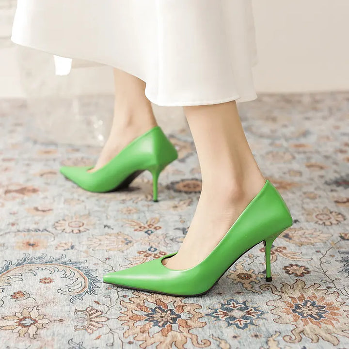 Vibrant green Qanari stiletto heels crafted from high-gloss faux leather, featuring a sharp pointed toe, low-cut vamp, and a distinctive sculpted heel base, shown on a model&