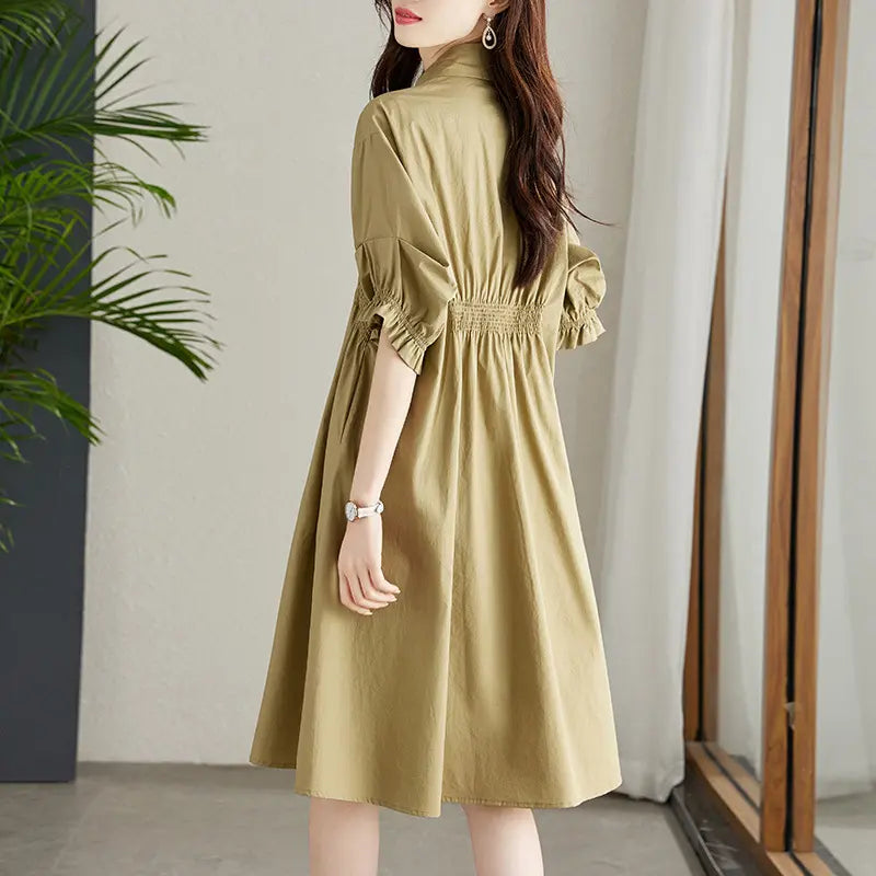 A woman models a Qelios midi shirt dress in a soft khaki color, showcasing the shirred waist, puff sleeves, and A-line skirt from a three-quarter rear view.