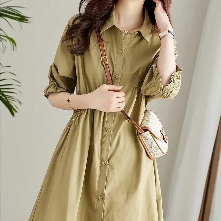A woman models a Qelios midi shirt dress in soft khaki, featuring a shirred waist, puffed sleeves with ruffled cuffs, and a classic button-front design.
