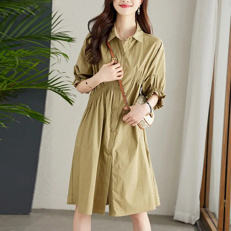 A woman wears a Qelios midi shirt dress in a khaki hue, featuring a shirred waist, puff sleeves with ruffled cuffs, and a button-front A-line skirt, styled casually.