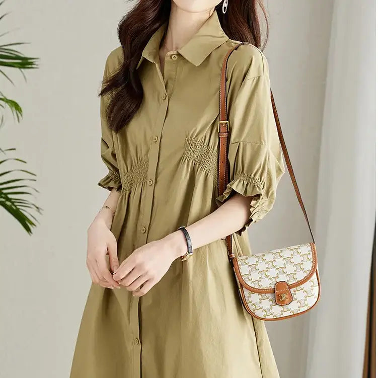 A smiling woman models a light khaki Qelios midi shirt dress with shirred waist and puff sleeves, accessorized with a crossbody bag.