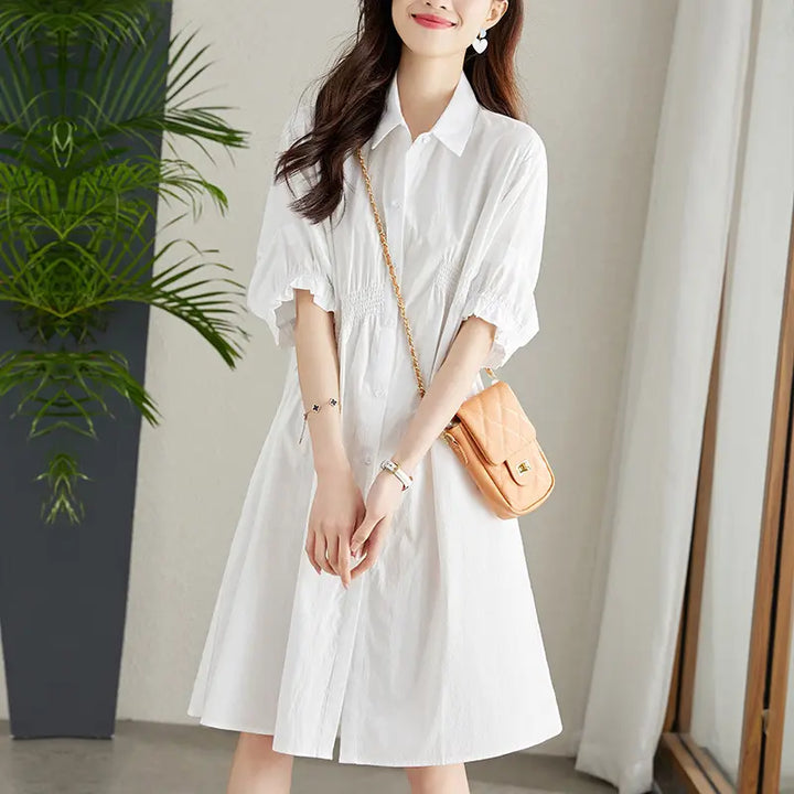 A woman wears a white Qelios midi shirt dress with shirred waist and puff sleeves, accessorized with a tan crossbody bag, presented in a lifestyle setting.