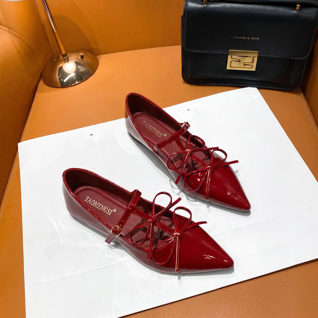 A pair of vibrant cherry red patent leather pointed-toe ballet flats with delicate bow details and adjustable ankle straps, showcased with a black handbag on a stylish couch.