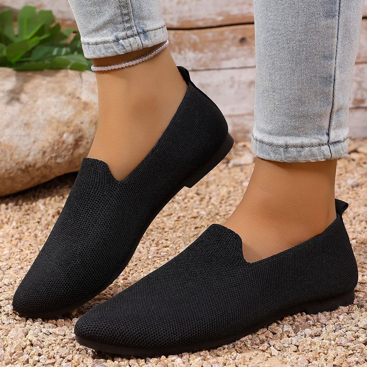 Black Quoris breathable knitted pointed toe flats are shown worn by a model on a textured pebble ground, showcasing their sleek design and comfortable knit upper.