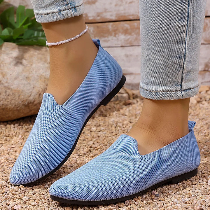 A pair of sky blue Quoris breathable knitted pointed toe flats are worn by a woman wearing light-wash jeans, styled in an outdoor setting on a pebble ground.