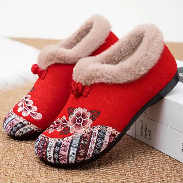 Red embroidered mules with plush faux fur lining and decorative bow accents, showcasing intricate floral and geometric patterns on the upper. These slip-on slippers offer cozy winter comfort.