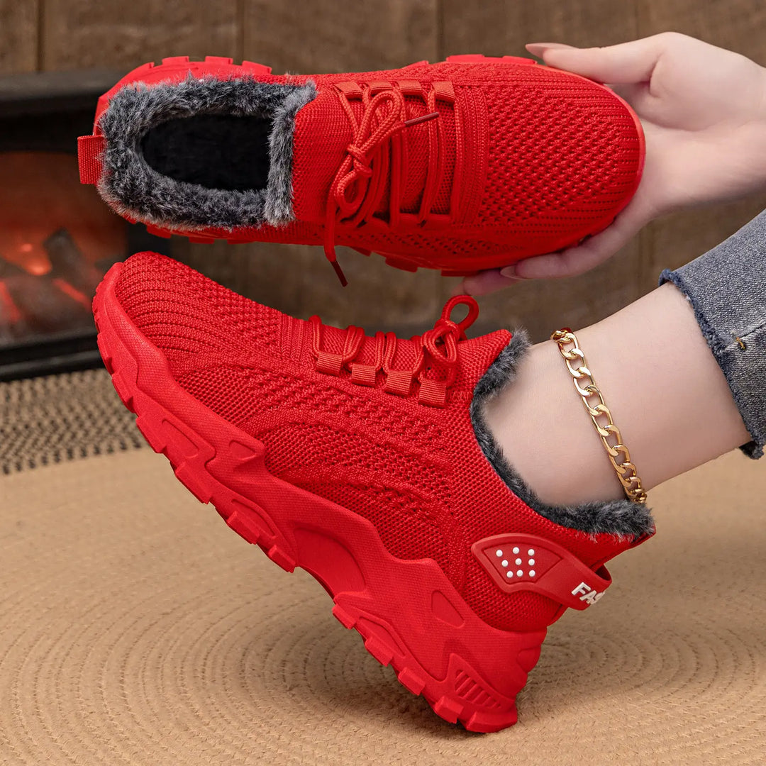 Red knit ankle boots with plush faux fur lining, shown on a foot with a gold anklet and another pair held in hand, against a warm, textured background.