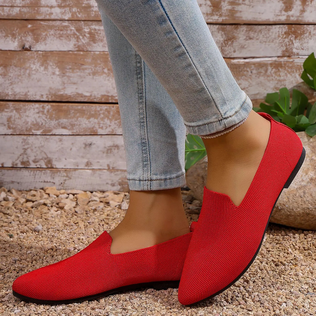 Red knitted pointed toe flats worn with light wash jeans, showcasing their sleek silhouette and breathable fly-knit texture on a pebble-strewn ground.