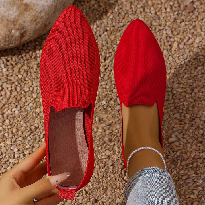 Red knitted pointed toe flats shown on a pebble ground, with one shoe held by a hand and the other on a foot adorned with an anklet.