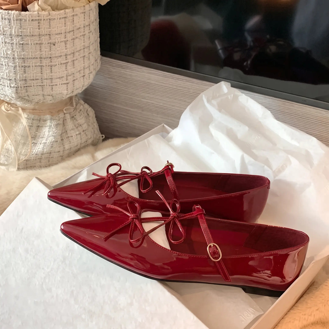 A pair of vibrant cherry red patent leather pointed-toe flats with delicate bows and thin adjustable ankle straps, presented on white packaging.