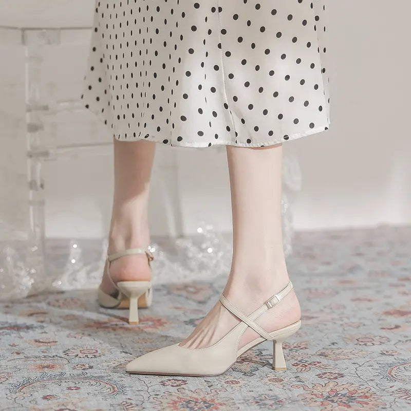 Elegant pointed-toe slingback heels in ivory, featuring a sculptural flared mid-heel and a delicate adjustable ankle strap, worn with a polka dot skirt on a patterned rug.