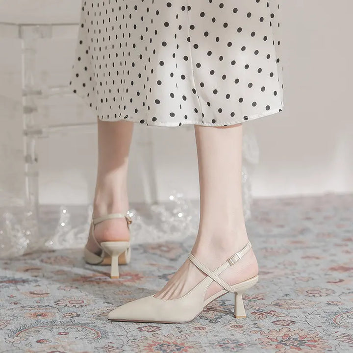 Elegant pointed-toe slingback heels in ivory, featuring a sculptural flared mid-heel and a delicate adjustable ankle strap, worn with a polka dot skirt on a patterned rug.