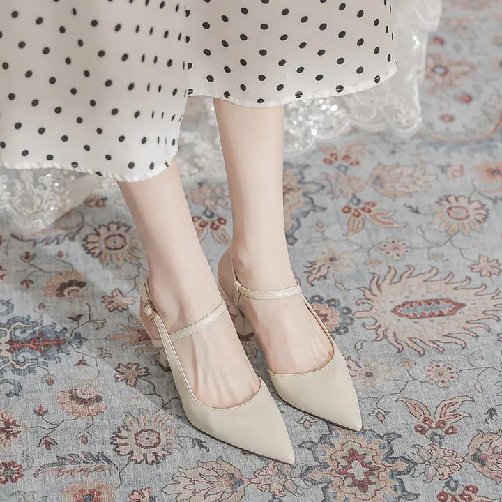 Elegant ivory pointed-toe slingback heels with a sculptural flared mid-heel and thin adjustable ankle strap, shown on feet in a lifestyle setting.