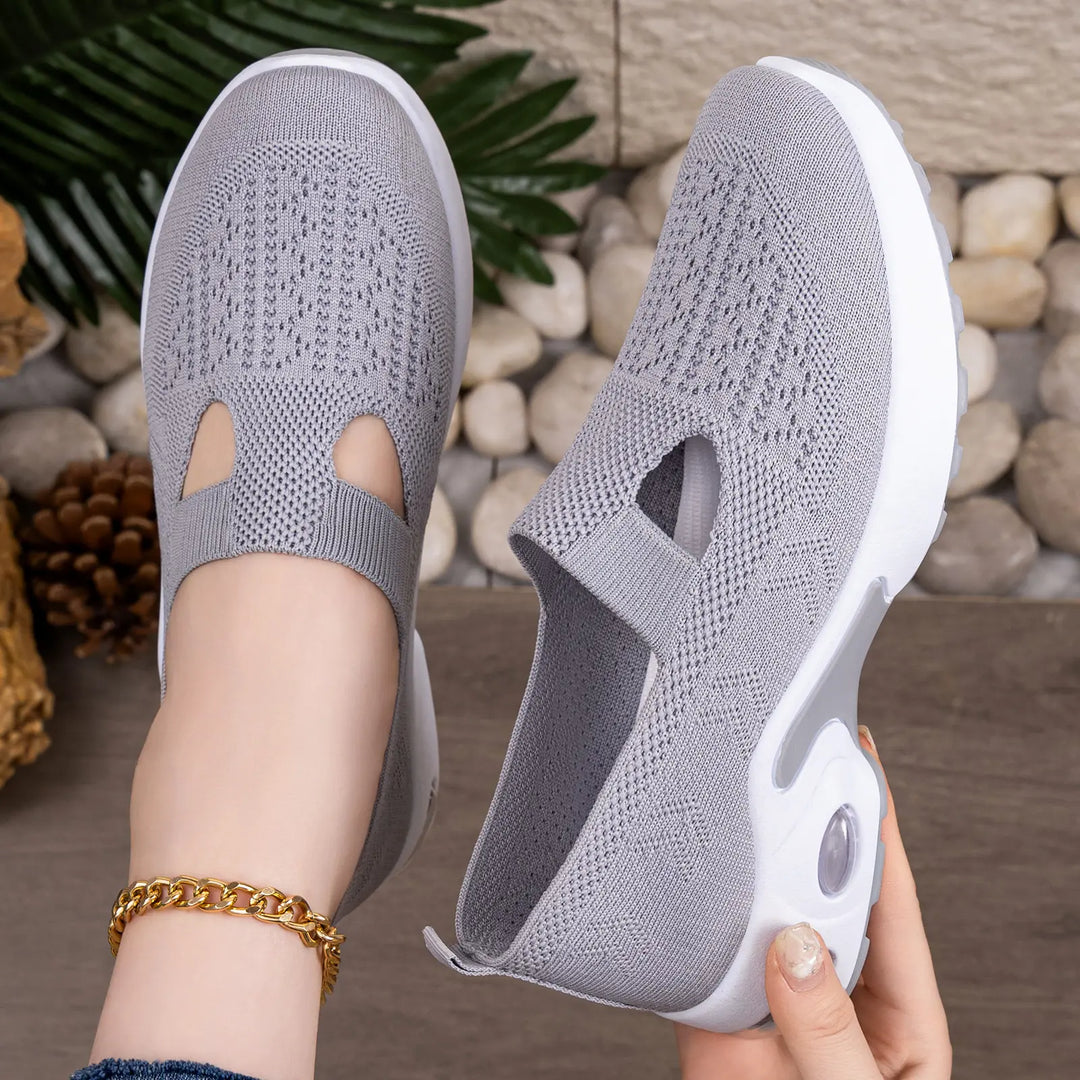 Grey Rilux breathable knit slip-on sneakers shown on a foot with a gold anklet and being held by a hand, featuring a visible air cushion sole and geometric knit pattern.