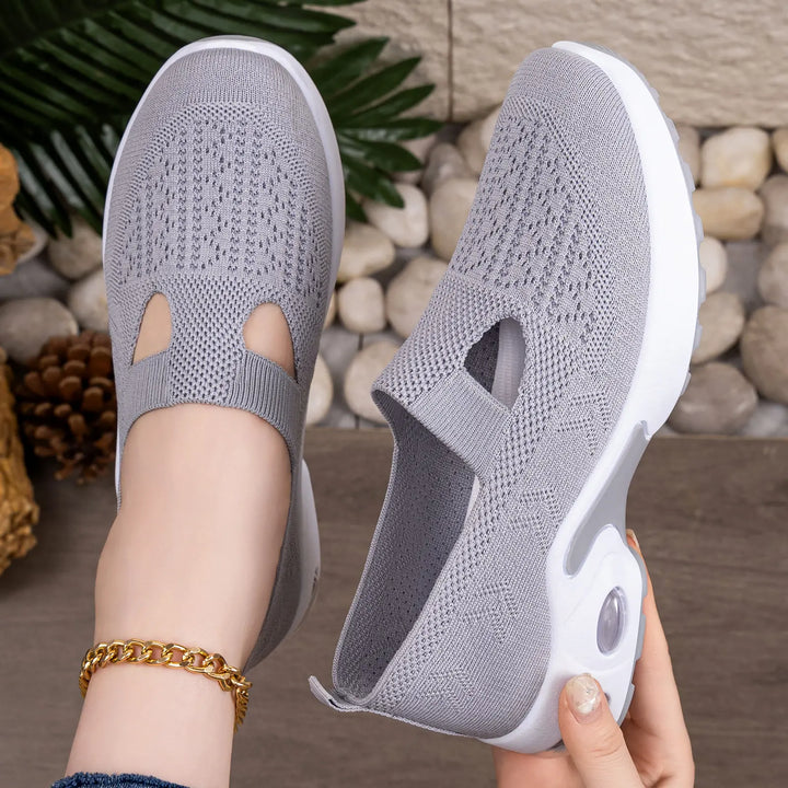 Grey Rilux breathable knit slip-on sneakers shown on a foot with a gold anklet and being held by a hand, featuring a visible air cushion sole and geometric knit pattern.