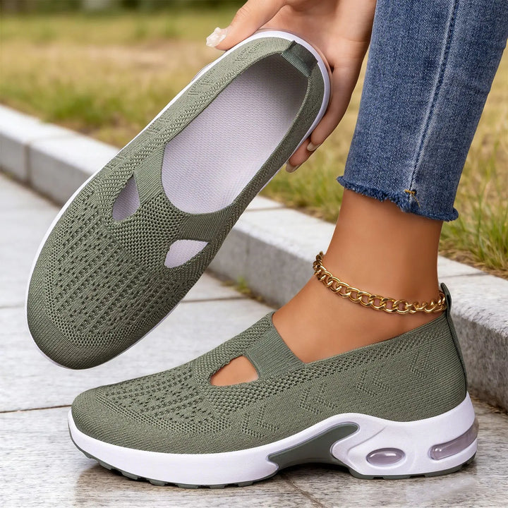 Sage green Rilux breathable knit slip-on sneakers worn by a woman, showcasing the air cushion sole and geometric knit pattern. A second shoe is held in hand against a blurred outdoor background.
