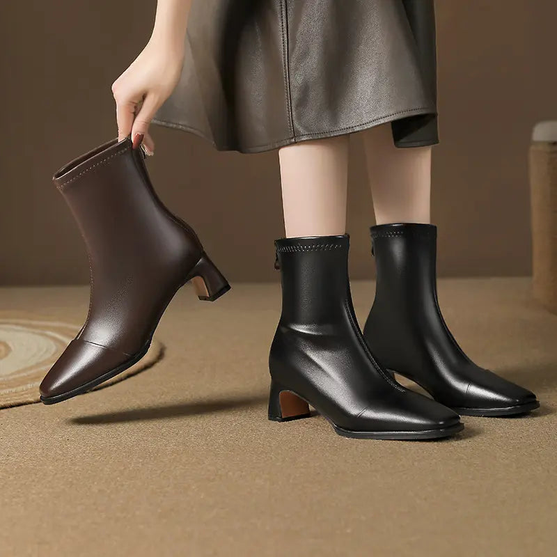 Model holding a brown square toe ankle boot next to a pair of black ones on foot, showcasing the block heel and mid-calf shaft.