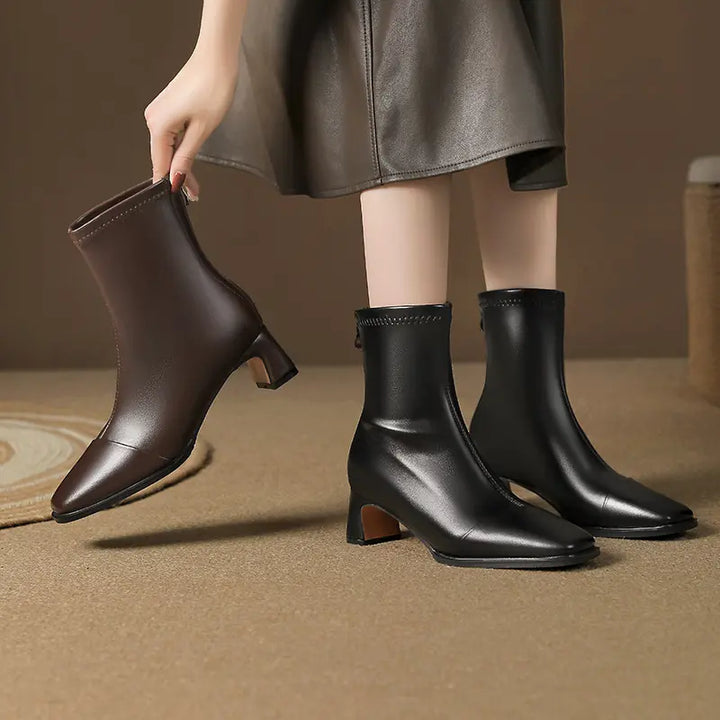 Model holding a brown square toe ankle boot next to a pair of black ones on foot, showcasing the block heel and mid-calf shaft.