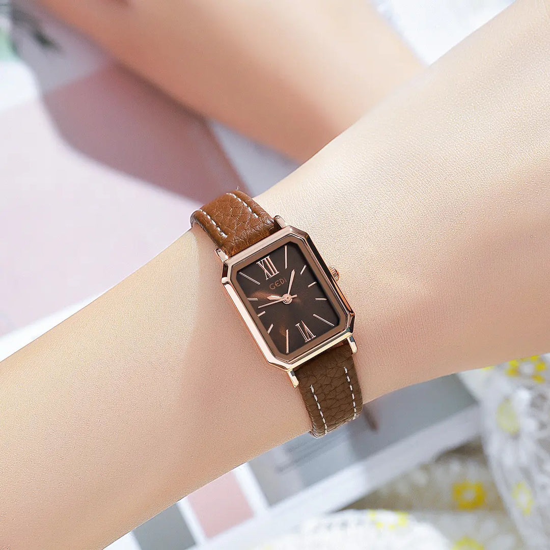A rose gold rectangular watch with a brown dial and Roman numeral markers is worn on a wrist, featuring a brown faux leather strap with white contrast stitching.