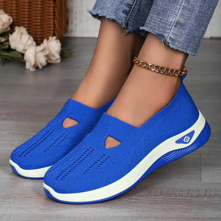 A pair of vibrant royal blue breathable knit slip-on sneakers worn by a woman, featuring a geometric weave pattern, white midsole, and a subtle ankle bracelet.