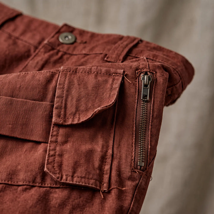 Close-up of rust-colored cotton twill cargo pants, showing matte fabric texture, button closure, a flap cargo pocket, and a side zipper detail.