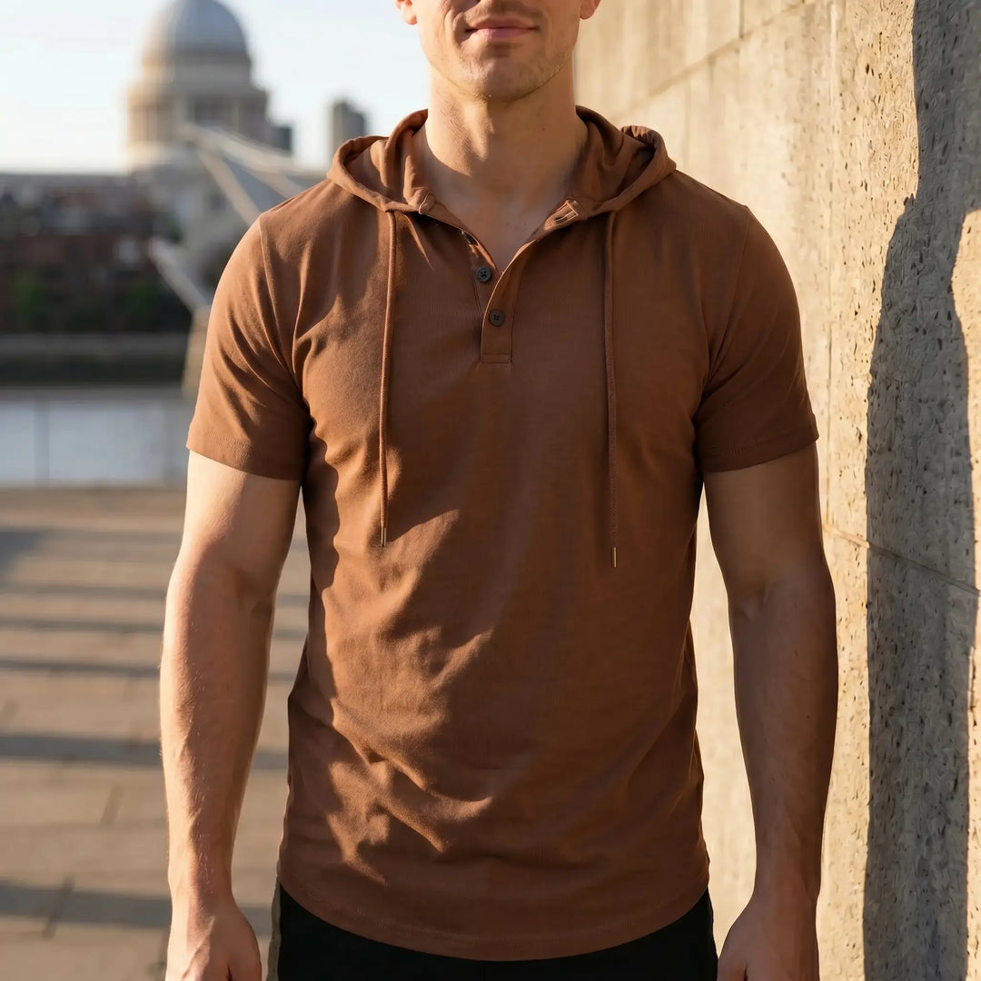 A man wearing a short-sleeve, earthy brown hooded henley tee stands outdoors, leaning against a textured wall with a blurred cityscape in the background. The tee features matte black buttons and drawstrings.