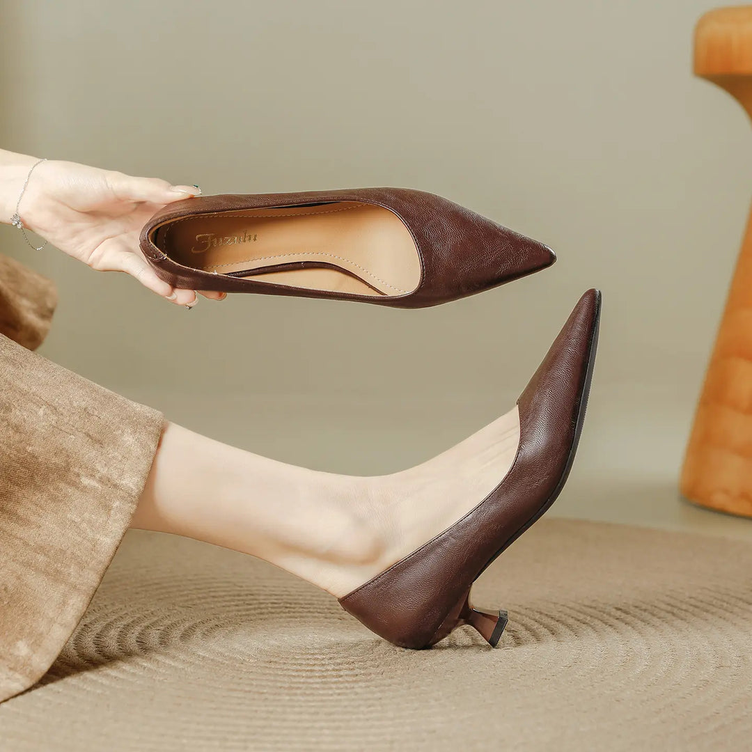A pair of women's brown faux leather pointed-toe pumps with a flared kitten heel, shown on a foot and held by a hand. This lifestyle image showcases the shoe's sophisticated silhouette and mid-height heel.
