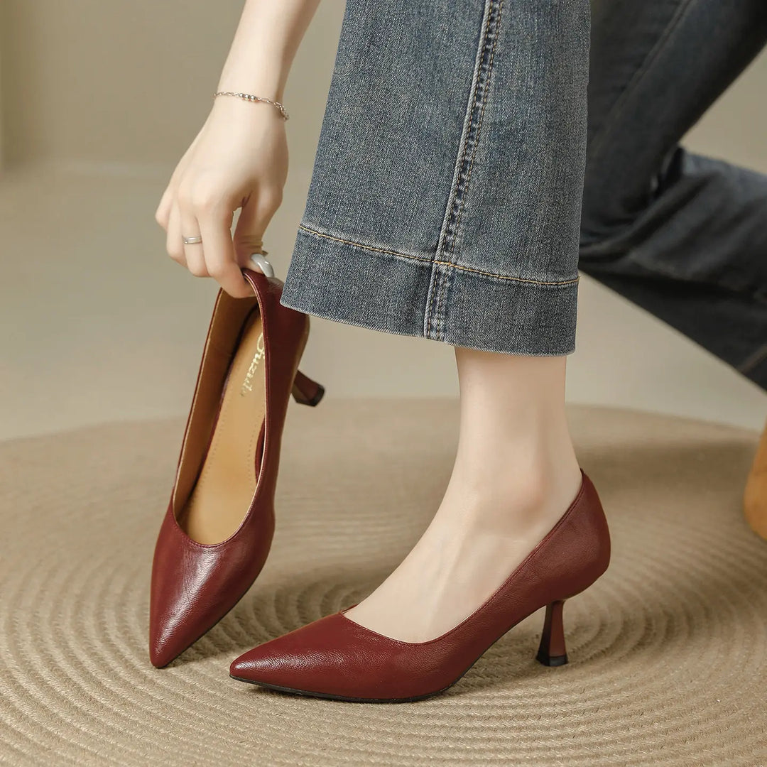 A pair of burgundy faux leather pointed-toe pumps with a sculpted flared kitten heel, shown from a three-quarter angle with one shoe on a foot and the other held.