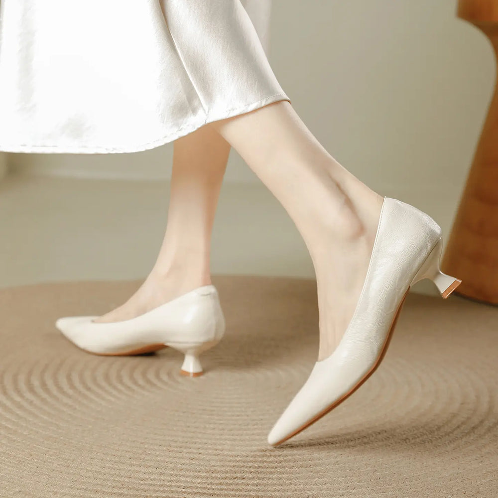 Elegant cream-colored women's pointed-toe pumps with a distinctive flared kitten heel, crafted from smooth faux leather, shown from a three-quarter angle on a textured mat.
