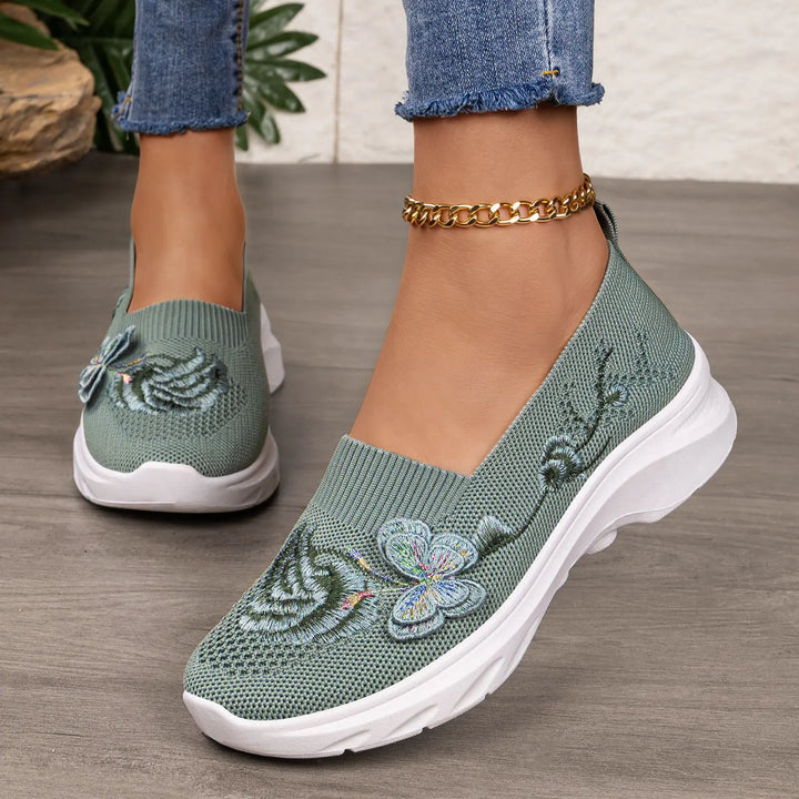 Close-up of sage green embroidered knit slip-on sneakers with floral and butterfly motifs, worn on a woman&