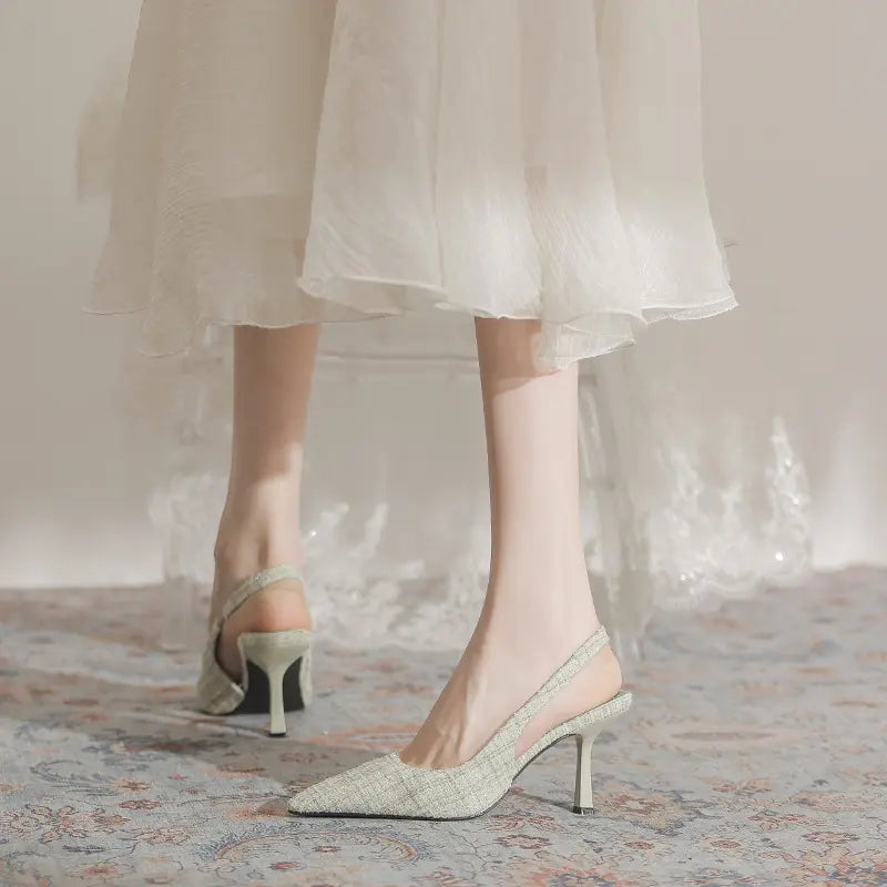 Elegant sage green pointed-toe slingback heels with textured tweed-like fabric and a slender stiletto heel, worn by a model with a flowing white dress.