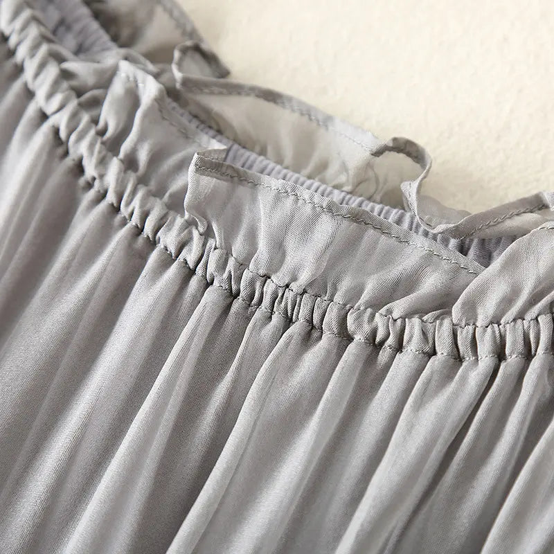 Close-up view of a muted grey pure silk slip dress, showcasing the delicate ruffled neckline, gathered elasticated waist detail, and flowing sheer fabric texture.