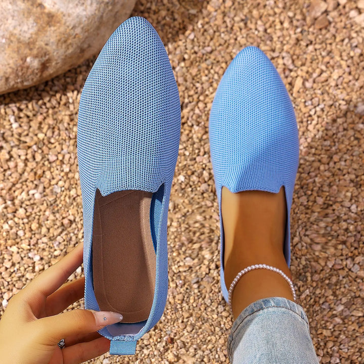 A pair of sky blue knitted pointed toe flats shown from a top-down perspective on a beach-like ground, one shoe on a foot with jeans and an anklet, the other held by a hand.