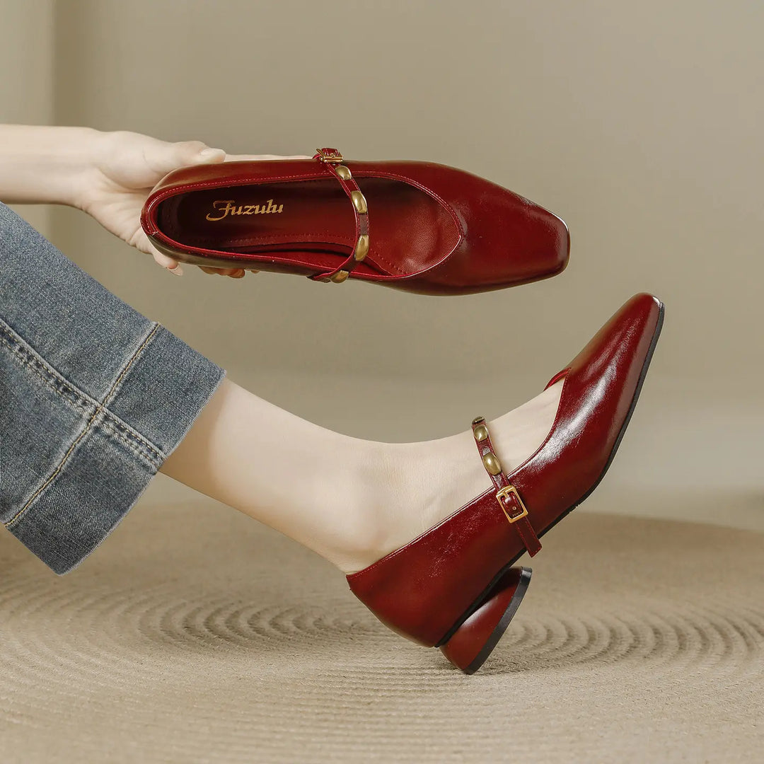 A pair of deep burgundy wine square toe Mary Jane shoes with a low block heel and a gold-tone buckle strap adorned with studs. One shoe is worn on a foot, showcasing the side profile and cut, while the other is held in hand to display the interior.