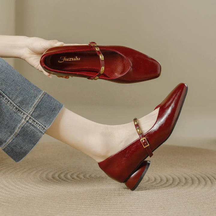 A pair of deep burgundy wine square toe Mary Jane shoes with a low block heel and a gold-tone buckle strap adorned with studs. One shoe is worn on a foot, showcasing the side profile and cut, while the other is held in hand to display the interior.