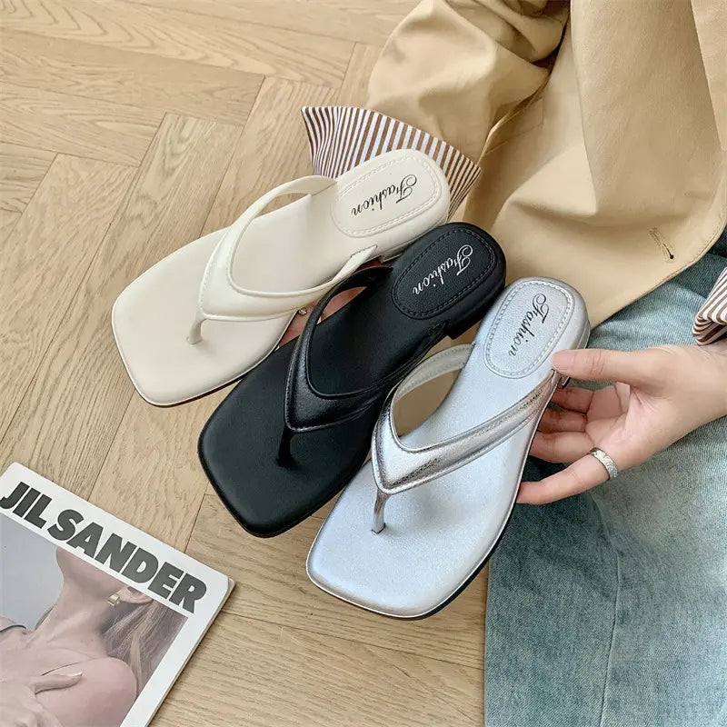 Three pairs of chic square-toe thong sandals in white, black, and metallic silver, laid out on a textured surface next to a magazine.