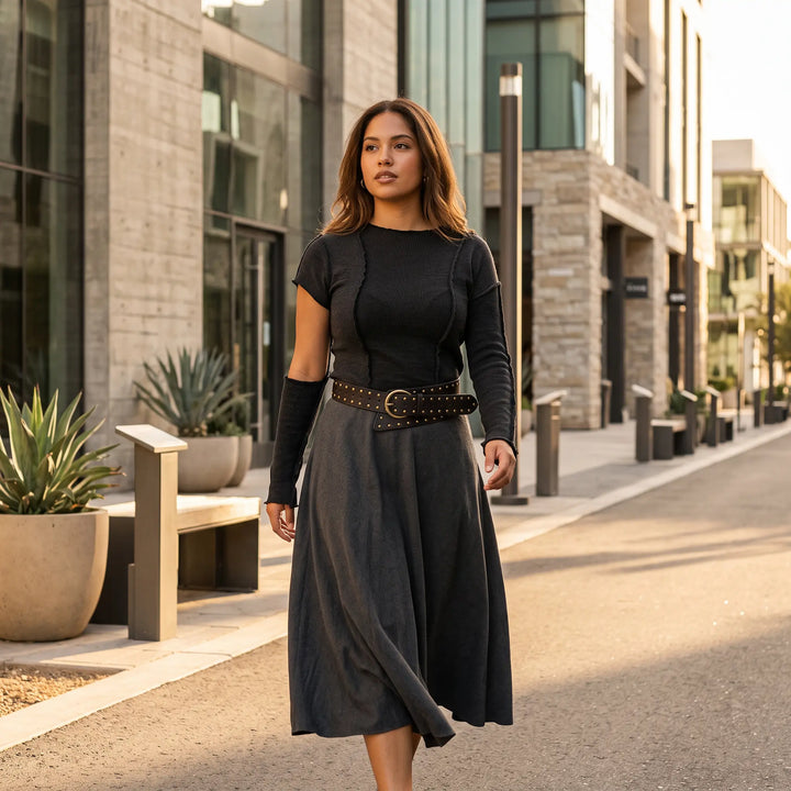 Woman wearing a wide black PU leather studded belt with antique brass hardware cinching a dark grey midi skirt and black top, walking on an urban street during golden hour.
