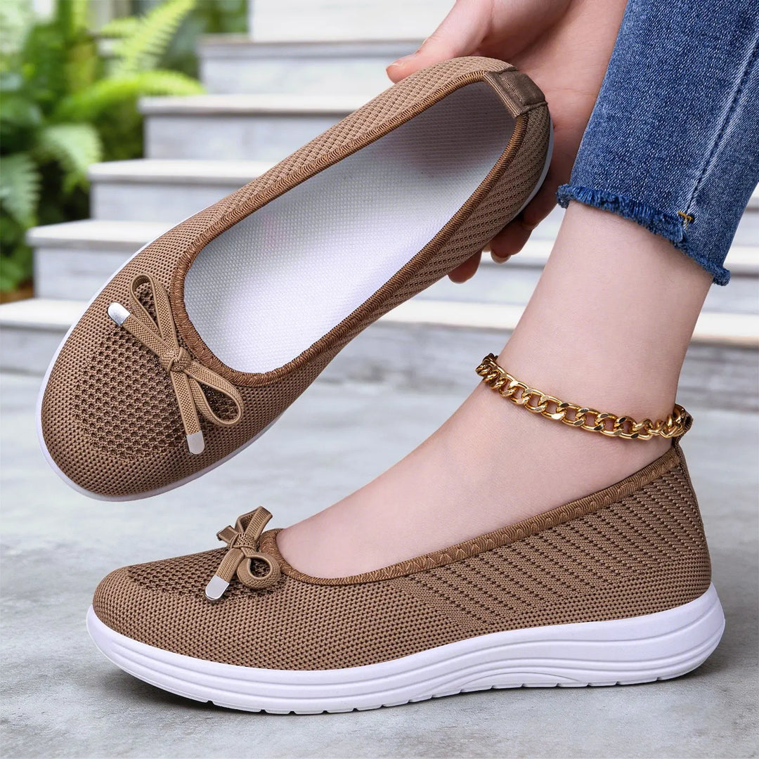 Women's breathable knit slip-on flats in taupe, featuring a decorative bow and a comfortable white EVA sole, shown styled on a foot with denim jeans and a gold ankle bracelet.