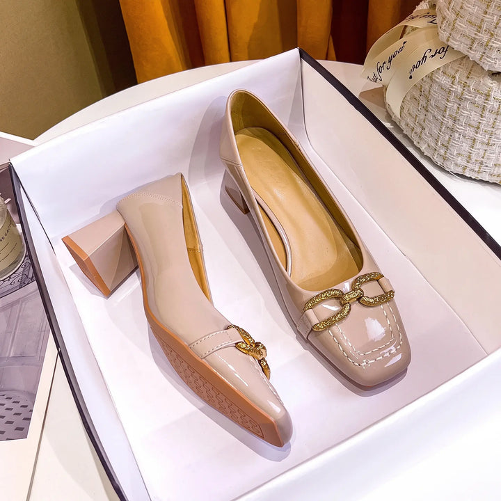 Pair of sophisticated taupe patent leather pumps with a square toe and decorative gold-tone buckle embellishment, displayed in a studio setting.